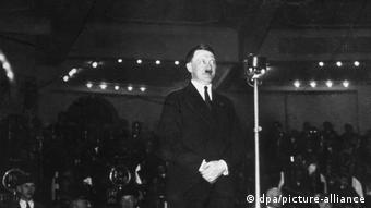Adolf Hitler steht vor einem Mirkofon, hinter ihm sitzen viele Menschen 