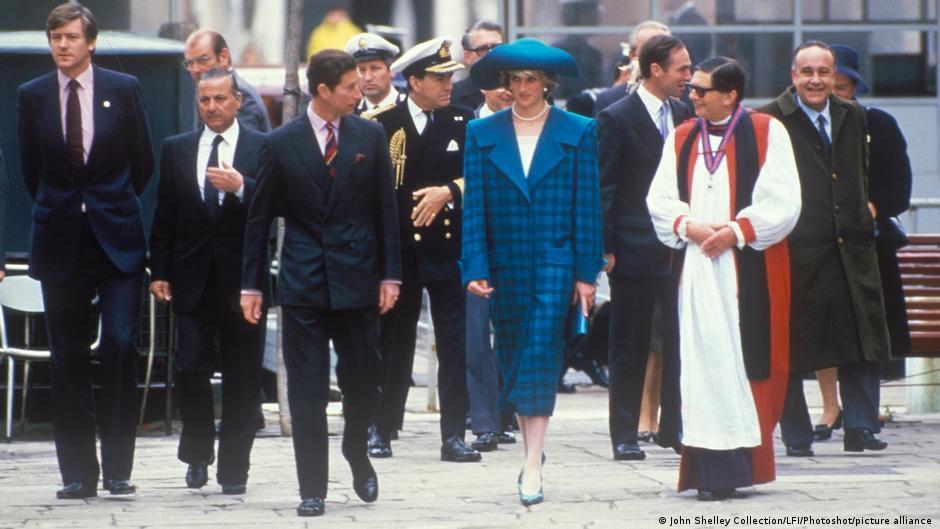 Diana beim Spaziergang in einer Gruppe von Männern in dunklen Anzügen und Marineuniformen. Neben ihr geht ein kirchlicher Würdenträger in einem weiß-rot-schwarzen Gewand.