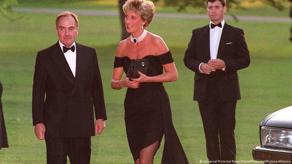 Lady Diana mit einem knappen schwarzen Cocktailkleid mit Chiffonschal, in den Händen eine Clutch, neben ihr zwei Herren mit Smoking.