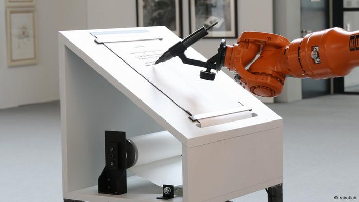 Ein Roboterarm schreibt mit einem Stift einen Text auf ein Papier, das auf einem querstehenden Tisch befestigt ist