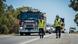 Two men in reflective vests walk down a road in Australia with emergency vehicles following Two men in reflective vests walk down a road in Australia with emergency vehicles following