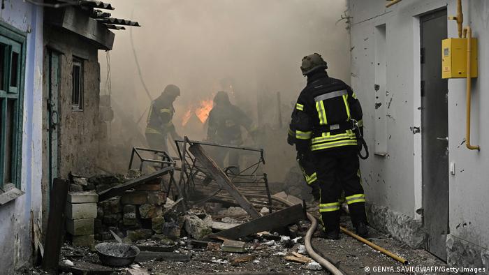 Drei Feuerwehrmänner gehen durch zerstörtes Haus, im Hintergrund glimmt noch ein Feuer