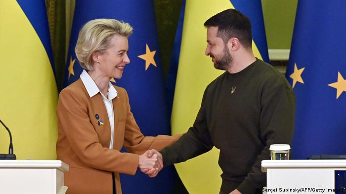 Mujer rubia de traje da la mano a hombre con barba. Detrás, banderas de la UE y de Ucrania.