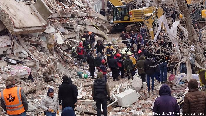 Menschen stehen auf Trümmern zusammengestürzter Häuser, einige Männer graben 
