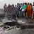 Dutzende Menschen stehen am Montag (07.03.2011) in Abodo vor einem Haufen Asche, der in Teilen noch brennt