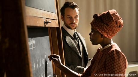 Filszene eine Herero-Frau löst mathematische Aufgaben an einer Tafel in einem Vorlesungssaal. Dabei wird sie von einem Mann, dem Ethnologen Alexander Hoffmann, angeschaut