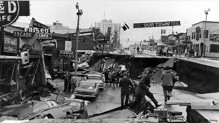 Schwarzweißaufnahme: Menschen laufen neben eingebrochenen Autos über eine abgesackte Straße (Anchorage, USA) nach dem Erdbeben in Alaska 1965