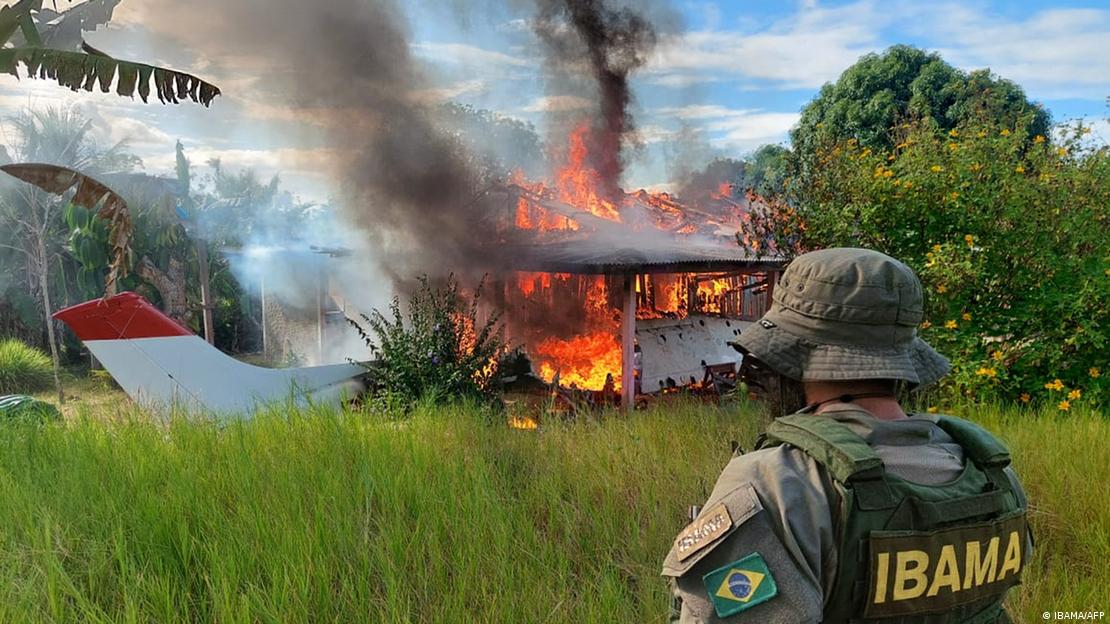 Pessoa de costas, com colete do Ibama, observa avião de pequeno porte pegando fogo em área com mata