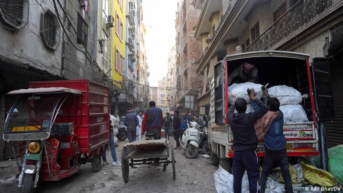 Elektroschrott-Sammler beladen auf einer engen Gasse in Neu Delhis Vorort Seelampur kleine Lasten-Motorräder mit Säcken, andere Menschen fahren mit Lastenrädern oder gehen zu Fuß durch die Straße