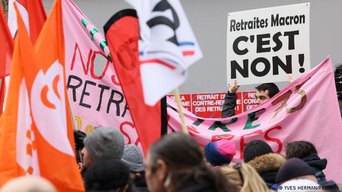Menschen stehen mit orange-, rosafarbenen und weißen Fahnen so wie Transparenten auf einer Straßen in Paris und demonstrieren gegen die geplante Rentenreform der Regierung