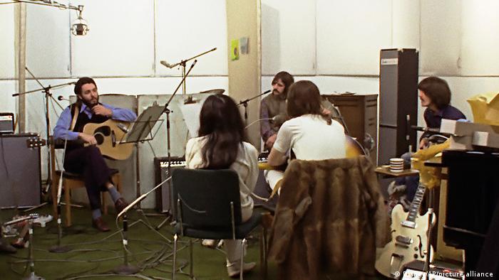 Die vier Beatles sitzen im Kreis im Studio und diskutieren über eine Aufnahme, im Kreis neben John Lennon sitzt Yoko Ono.