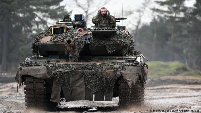 Leopard-2-Kampfpanzer fährt durch den Schlamm, in der Luke steht ein Mann in militärischer Tarnfleck-Kleidung 