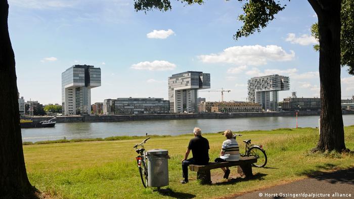 Two cyclists take a break on a bench and look at the Rhine, in the background the crane houses in Cologne, Germany