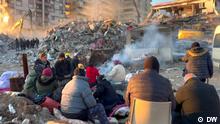 17.02.2023 A group of people is seen huddling around a fire against the backdrop of a collapsed building and heavy machinery at work in the rubble. 