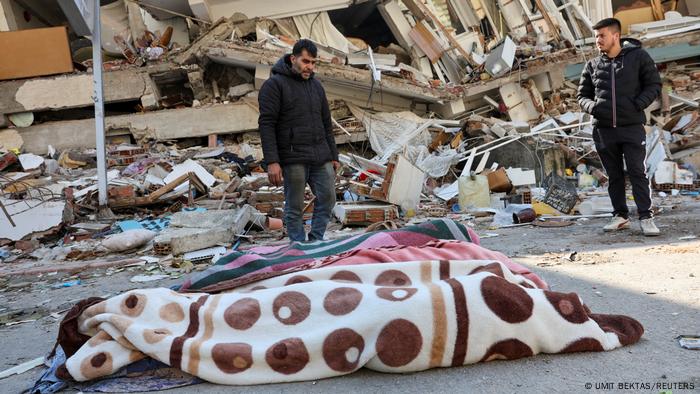 Los cuerpos de tres víctimas, envueltos en mantas, frente a una casa derrumbada.