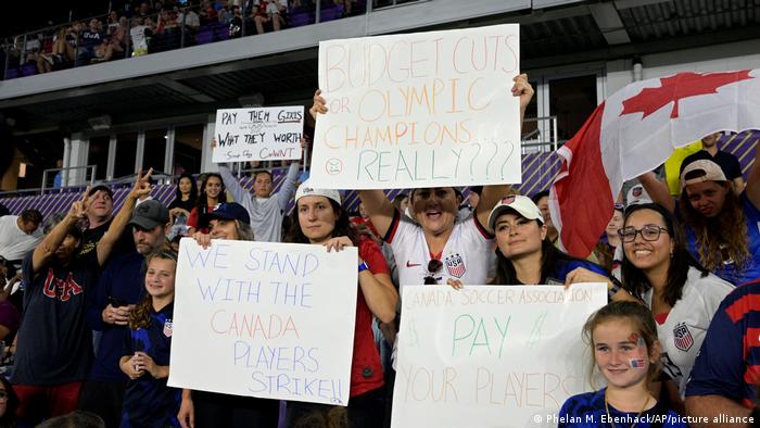Fans in Orlando halten Transparente hoch, auf denen sie die Forderungen der kanadischen Fußballerinnen unterstützen. 