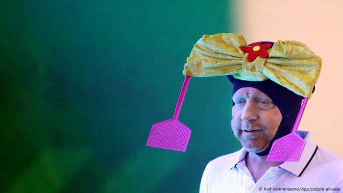 Boris Becker trägt in einer Fernsehshow eine Mütze, an der zwei Fliegenklappen befestigt sind.