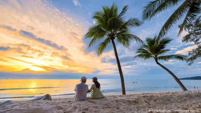 Ein Pärchen sitzt am Strand von Phuket neben Palmen und genießt den Sonnenuntergang.