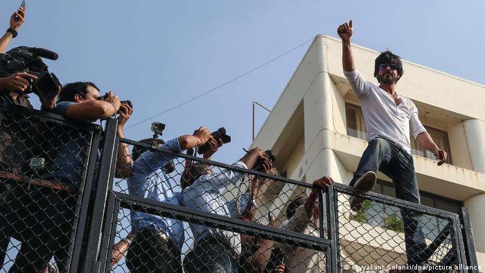 Schauspieler Shah Rukh Khan winkt auf einem Gitter stehend seinen Fans zu. mehrere Foto- und Filmkameras sind auf ihn und die Menge, die nicht im Bild ist, gerichtet.