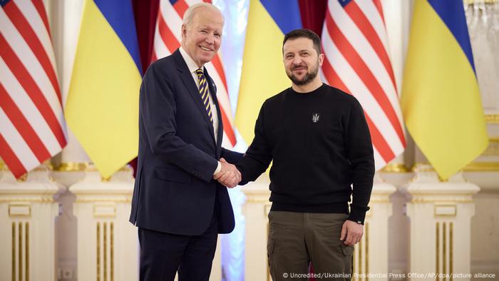 US President Joe Biden and Ukrainian President Volodymyr Zelenskyy shake hands and look towards cameras against a background of US and Ukrainian flags