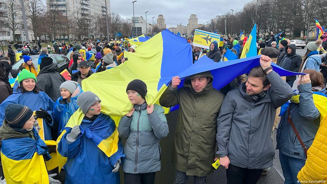A crowd of people marching through Berlin on February 24, 2023. They are lined up in a column, carrying a very large long Ukraine flag, which the people at the front of the line are also using to shield them from the rain. 