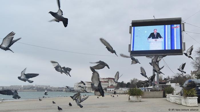 Tauben fliegen am Strand von Sewastopol auf der Krim vor einem Fernsehbildschirm, der den russischen Präsidenten Putin während seiner jährlichen Rede zur Lage der Nation zeigt