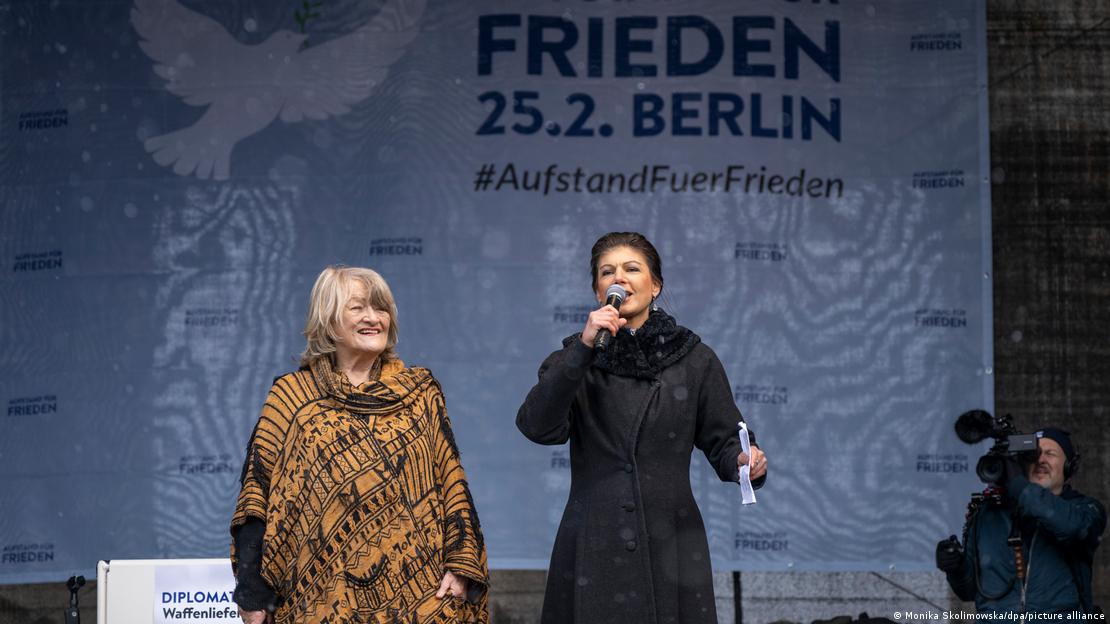 Die Frauenrechtlerin Alice Schwarzer (l.) und BSW-Chefin Sahra-Wagenknecht stehen nebeneinander auf der Bühne für die von ihr initiierte Demo am 25. Februar 2023 in Berlin. Das Motto ist neben einer weißen Friedenstaube auf einem riesigen weißen Transparent vor dem Brandenburger Tor in Form eines sogenannten Hashtags zu sehen: "#'AufstandFuerFrieden".   