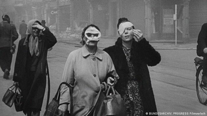 Zwei Menschen mit Verbänden im Gesicht schauen in die Kamera, während sie durch die Straßen Frankfurts laufen