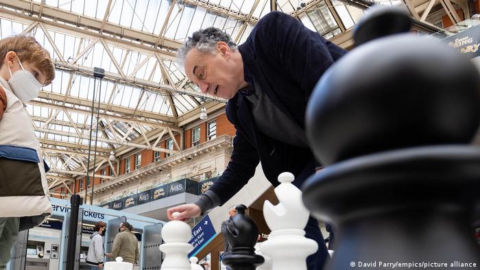 Malcolm Pein spielt in einer Bahnhofshalle mit riesigen Schachfiguren gegen ein Kind Schach