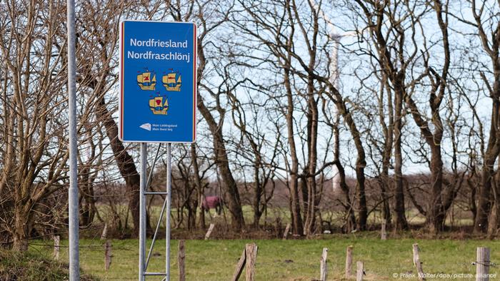 Ein blaues Straßenschild vor Bäumen und Feldern mit der Aufschrift 'Nordfriesland' und der friesischen Variante 'Nordfraschlönj'. Auf dem Straßenschild sind zudem gelbe Schiffe zu sehen.