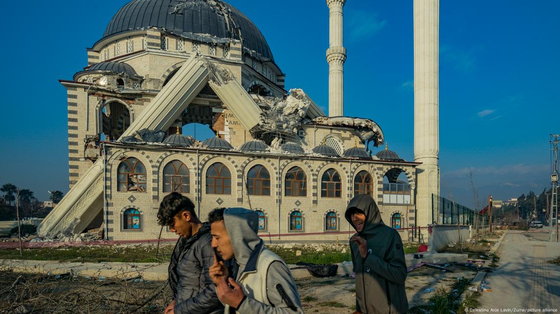Kahramanmaraş depremlerinin ardından Hatay'da ağır hasar gören, bir minaresi yıkılan bir cami ve önünde yürüyen üç genç - (20.02.2023)