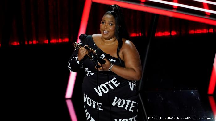 Lizzo steht bei einer Preisverleihung auf der Bühne und hält einen Award in der Hand. Auf ihrem schwarzen Kleid stehen weiße Schriftzüge mit dem Wort Vote (deutsch: wählen)