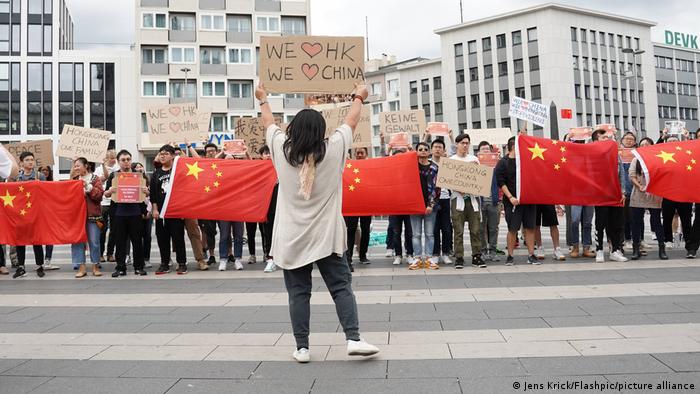 Junge Frau von hinten mit einem Plakat, auf dem die Botschaft We love Hongkong, we love China zu lesen ist, außerdem weitere Protestanten mit Plakaten und chinesischen Flaggen