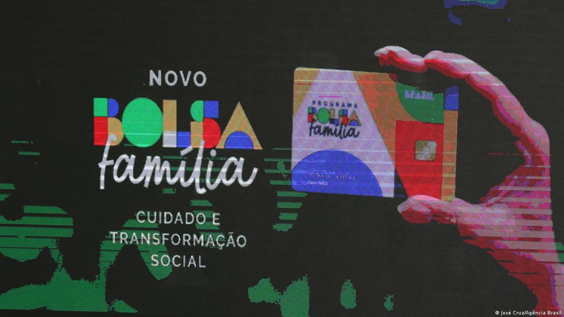 Em imagem projetada em telão, mão segura cartão do programa Bolsa Família com novo layout, que traz formas geométricas coloridas nas cores azul, amarelo, branco, verde e vermelho. 