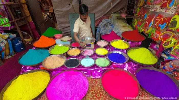 Ein indischer Verkäufer steht hinter einer Vielzahl an Tabletts, die Pulver in den verschiedensten Farben enthalten