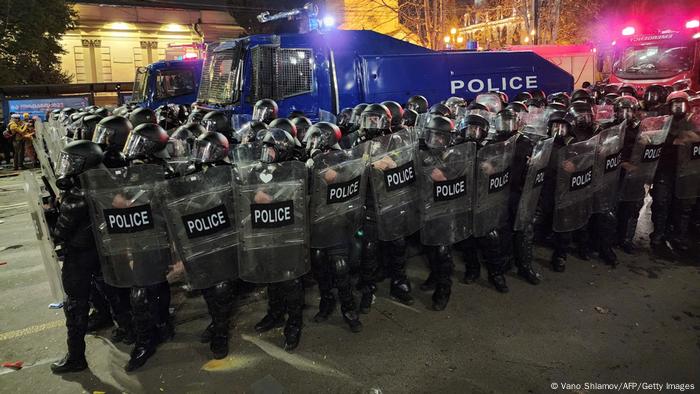 Einsatzpolizisten mit Plastikschilden und Helmen haben vor gepanzerten blauen Polizeifahrzeugen Aufstellung genommen