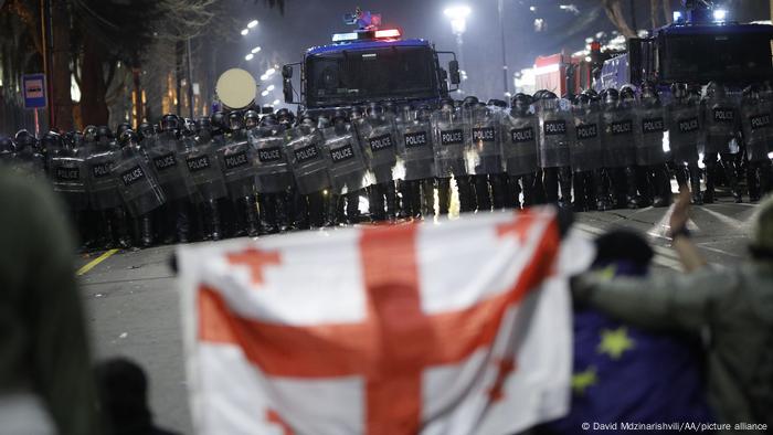 Una lína de policías con escudos y un cañón de agua al fondo, con manifestantes desenfocados en primer plano con banderas de Georgia y de la UE.
