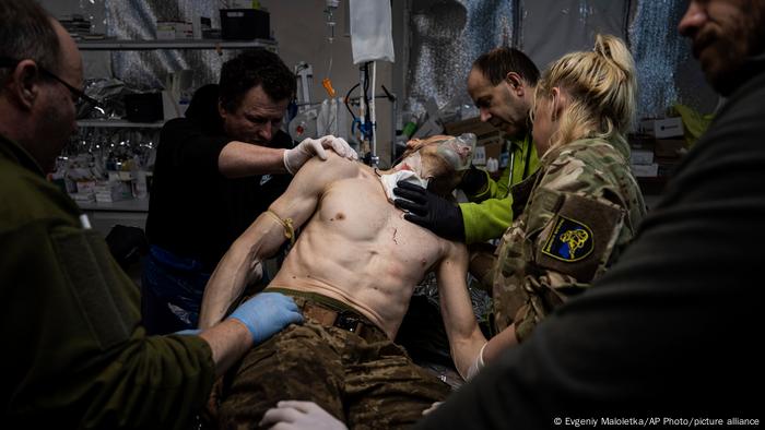 Un hombre con el torso desnudo, pantalones militares y máscara de oxígeno es atendido por cinco personas en una sala con equipos médicos. Una de ellas le pone una venda en el cuello.