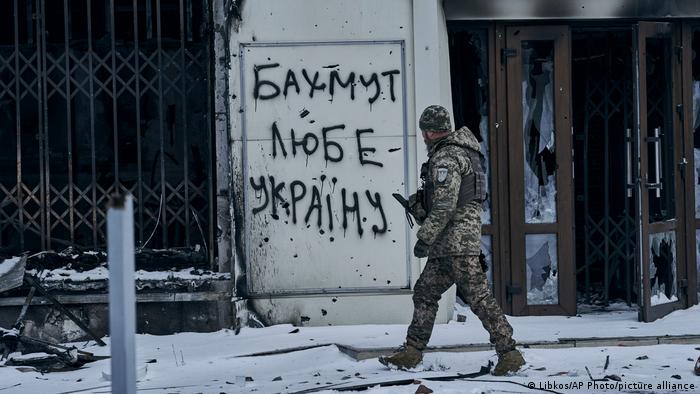 Un soldado ucraniano camina por la nieve junto a unas puertas de cristal destrozadas. Está mirando un grafiti garabateado en una pared, en el que se lee Bajmut ama a Ucrania en cirílico.