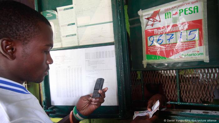  Kenia, Nairobi | Mobiles Bezahlsystem M-PESA: Mann nimmt Geld an M-PESA-Schalter entgegen, bedient Handy (Foto: Daniel Irungu/EPA/dpa/picture alliance)