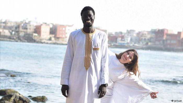 Ein Mann und eine Frau in weißen Gewändern stehen vor einem Gewässer, im Hintergrund ist eine Stadt zu sehen