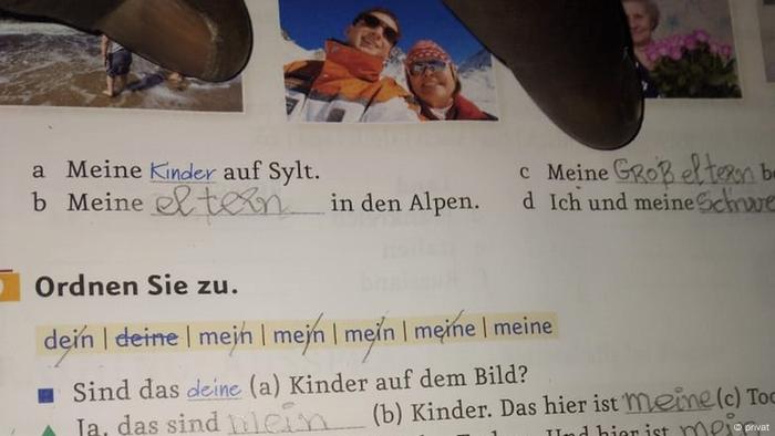 Blick in ein Aufgabenheft mit Fotos und Text, zwei Finger deuten auf die Aufgaben