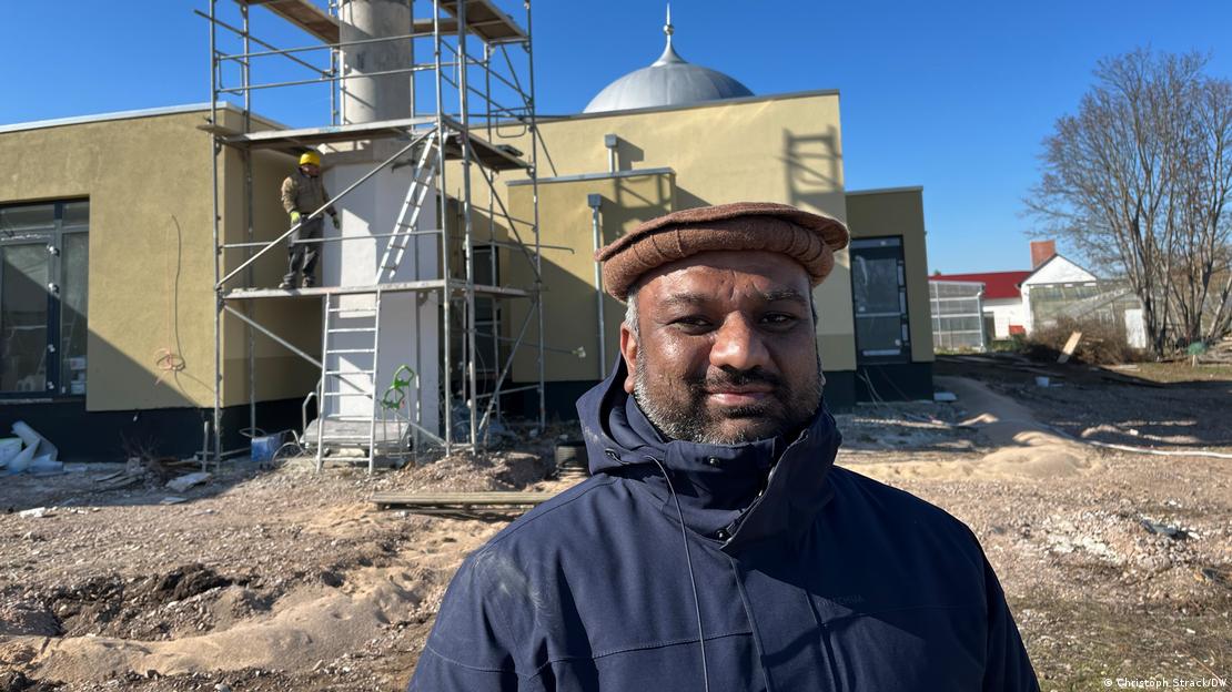 Malik stands in front of the construction site of the mosque wearing a hat and coat