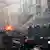 Italienische Polizisten stehen vor einem Gebäude in Kampfmontur. Ein Auto brennt derweil
