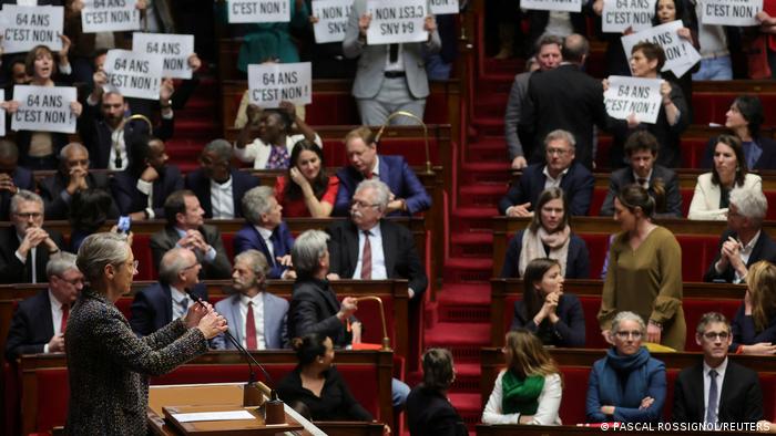 Menschen sitzen in einem Halbkreis und halten Schilder mit der Aufschrift 64 Ans C'est Non!. Vorne steht eine Frau vor einem Rednerpult.