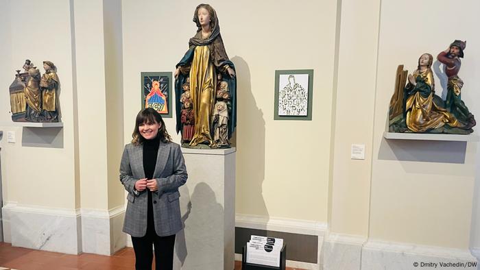 Eine Frau in einem grauen Blazer steht vor einer Madonnenstatue und zwei Gemälden. Rechts und links befinden sich weitere vergoldete Figurengruppen.