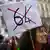 Εικόνα διαδηλώτριας που κρατάει πλακάτ που γράφει τον αριθμό 64 διαγεγραμμένο με ένα μεγάλο Χ.