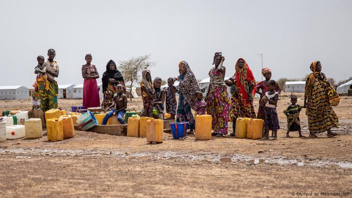 Eine Gruppe von Frauen und Kindern steht vor Plastikkanistern in einer trockenen Landschaft 