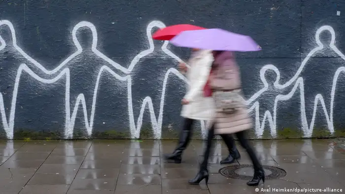 Women walking down the street with umbrellas, graffiti along a wall.