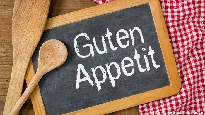 A chalkboard sign that says Guten Appetit, with wooden spoons to the side and a red-and-white tablecloth underneath.